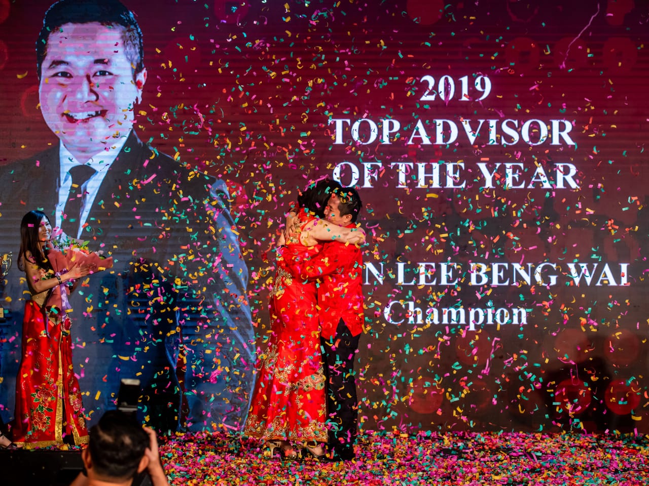 FLP Realty 新春晚宴舞台，彩纸纷飞，大屏幕写着 2019 TOP ADVISOR OF THE YEAR · Champion，Kevin 与人相拥。