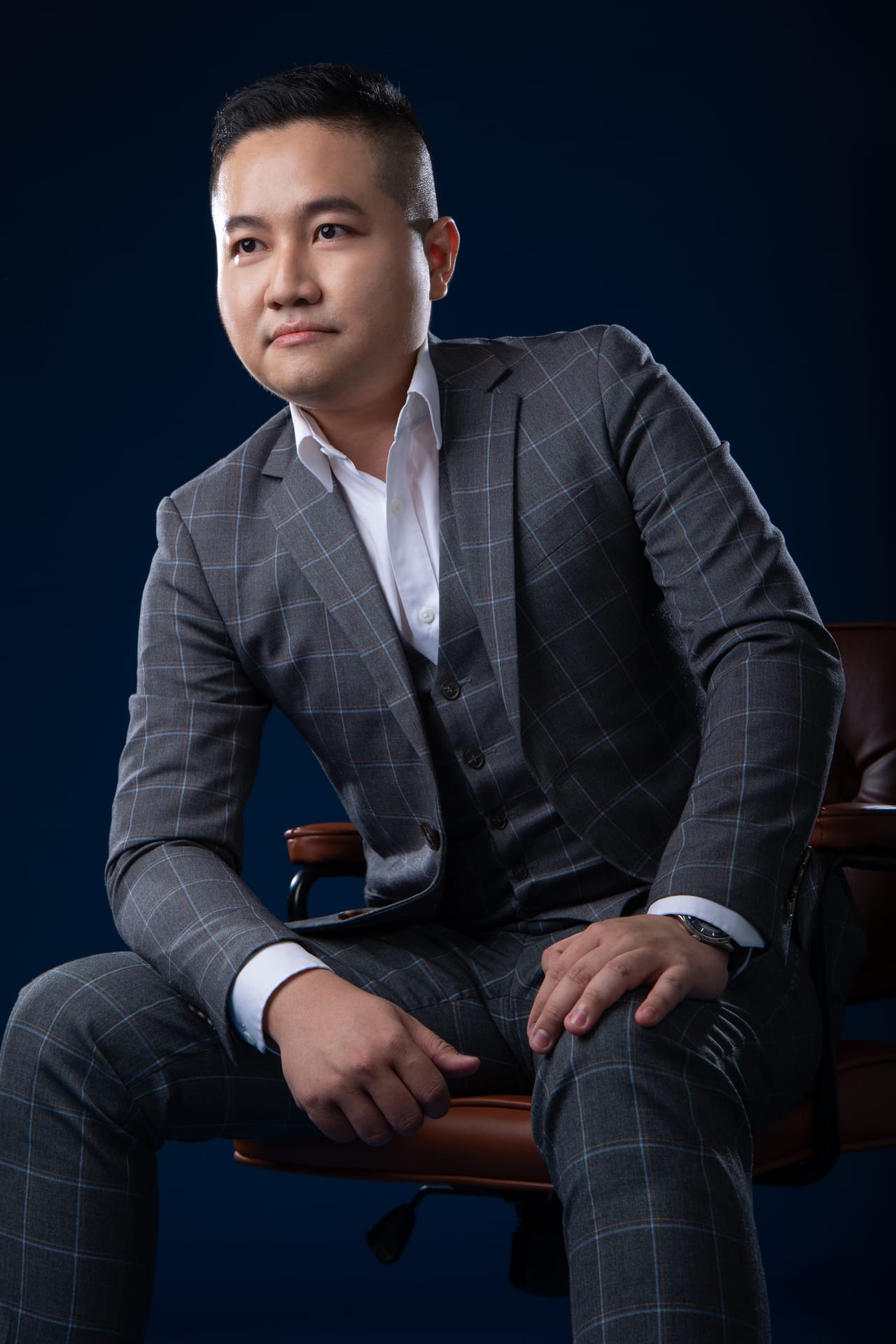 Against a deep-blue background, Kevin in a grey check suit sits leaning forward, elbows on his knees, looking at the camera.