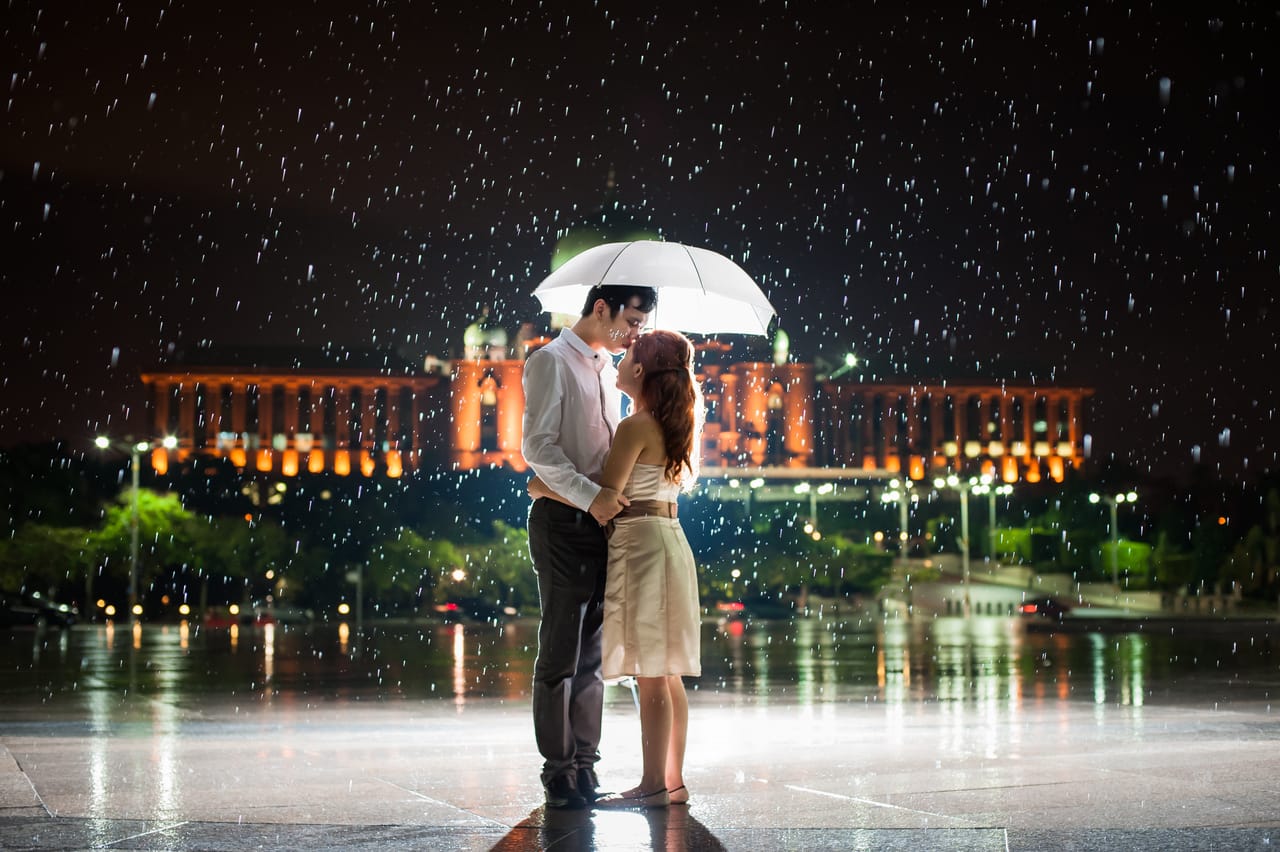 夜里下着大雨，一把白伞底下，两个人相拥，地面映着建筑的灯光。